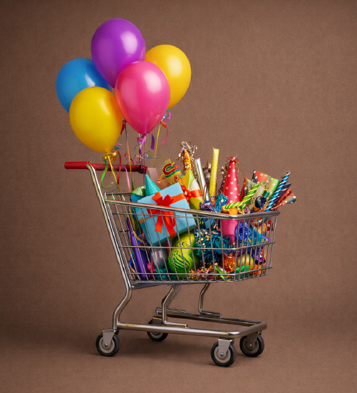 A silver metal shopping cart filled with colorful party supplies, including a light blue gift box with a red bow, several patterned party hats, and noisemakers. Five vibrant balloons in shades of blue, purple, pink, and yellow are tied to the handle and float above the cart against a solid brown background.