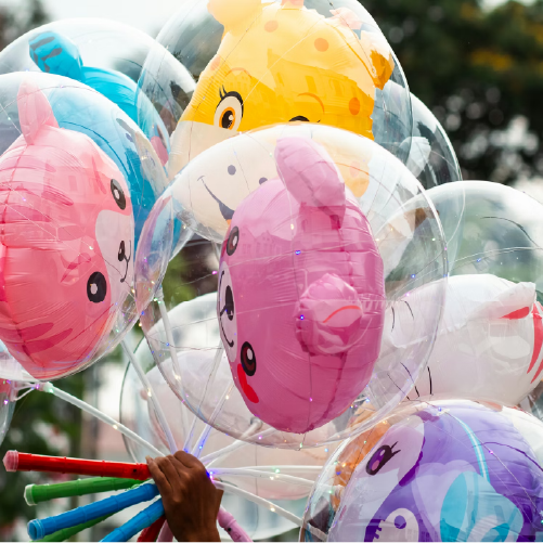 A hand holds a large bunch of glowing balloons against a blurred outdoor background. Each balloon consists of a large, clear outer sphere containing a smaller foil balloon shaped like a cute animal face—including a pink cat, a yellow giraffe, and a purple character. Tiny LED fairy lights sparkle inside the clear outer shells, and the balloons are mounted on vibrant red, blue, and green sticks.